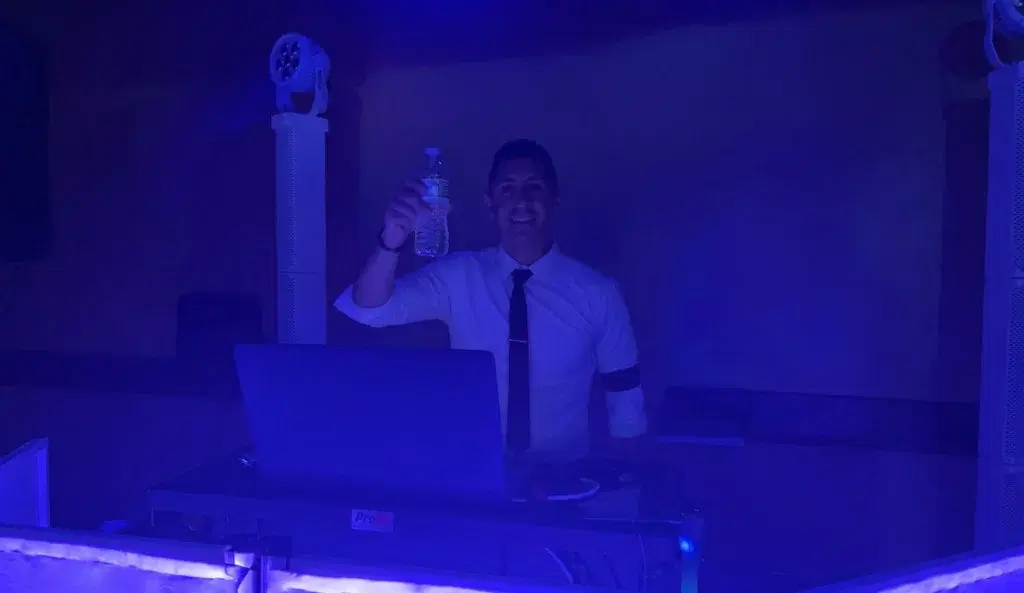 DJ NES smiling behind the booth in a white shirt and tie, holding up a water bottle under blue event lighting